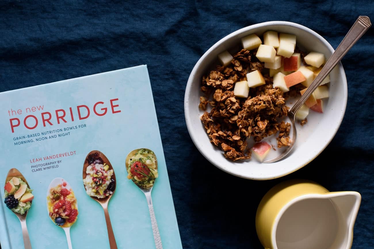 Tahini granola in a bowl on a blue tablecloth with a yellow milk pourer (recipe adapted from the cookbook, The New Porridge)