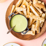 Baked yuca fries with cilantro dipping sauce on a wooden plate on a pink table.