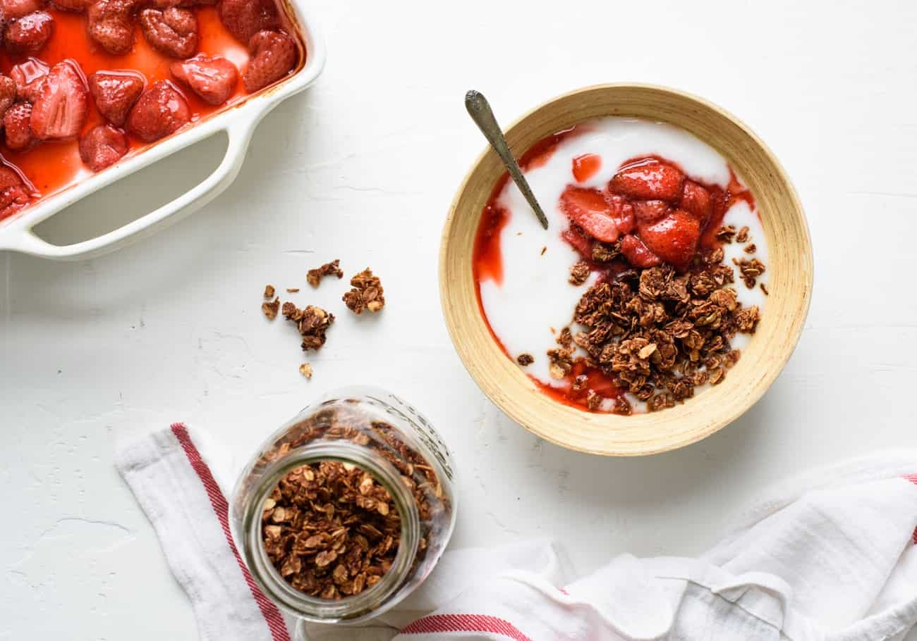 A bowl of vegan yogurt with chocolate coconut granola and roasted strawberries, alongside a mason jar of granola