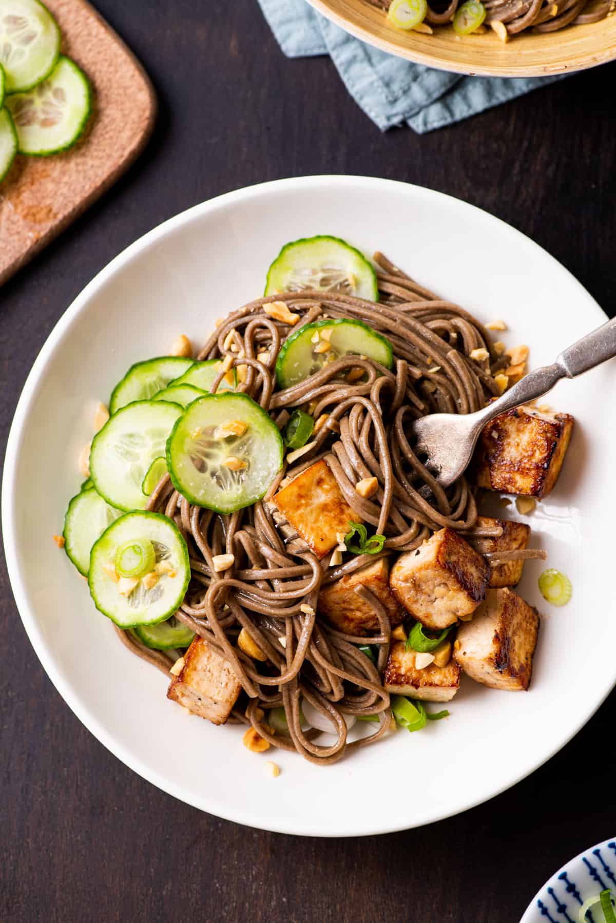 Soba noodle bowl with tofu, cucumbers, and peanuts on a dark wooden table.