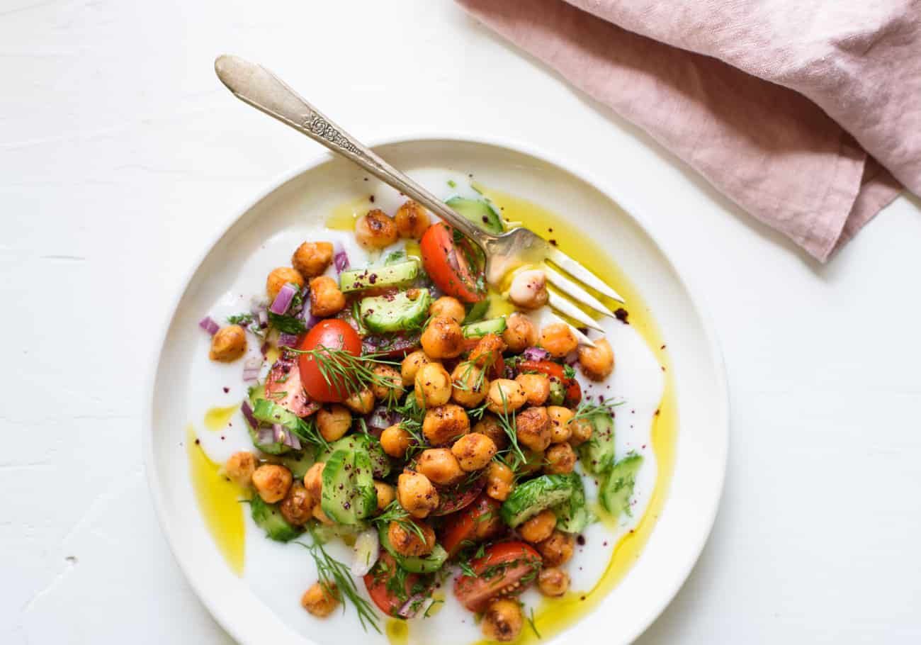 Savory yogurt bowl with chickpeas, cucumbers, tomatoes, and dill on a white plate next to a pink linen napkin