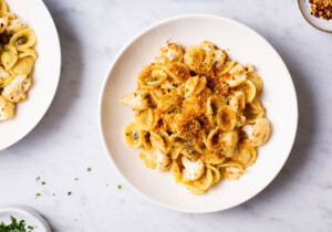 Lemony pasta with cauliflower and fried breadcrumbs in white bowls next to minced parsley.