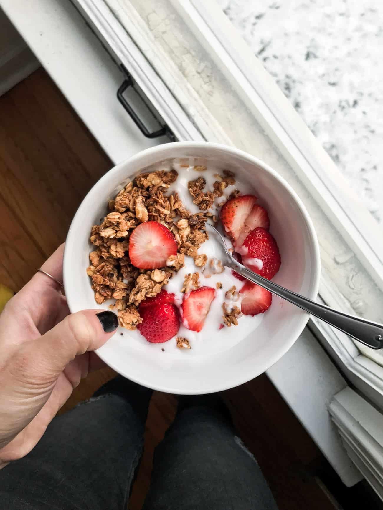 Woman's hand holding a white bowl of yogurt, granola and strawberries