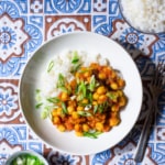 Bowl of chana masala on an Indian-patterned tablecloth next to rice and scallions