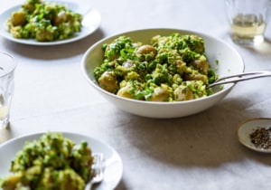 Bowl of smashed new potatoes with peas on a beige tablecloth next to white plates