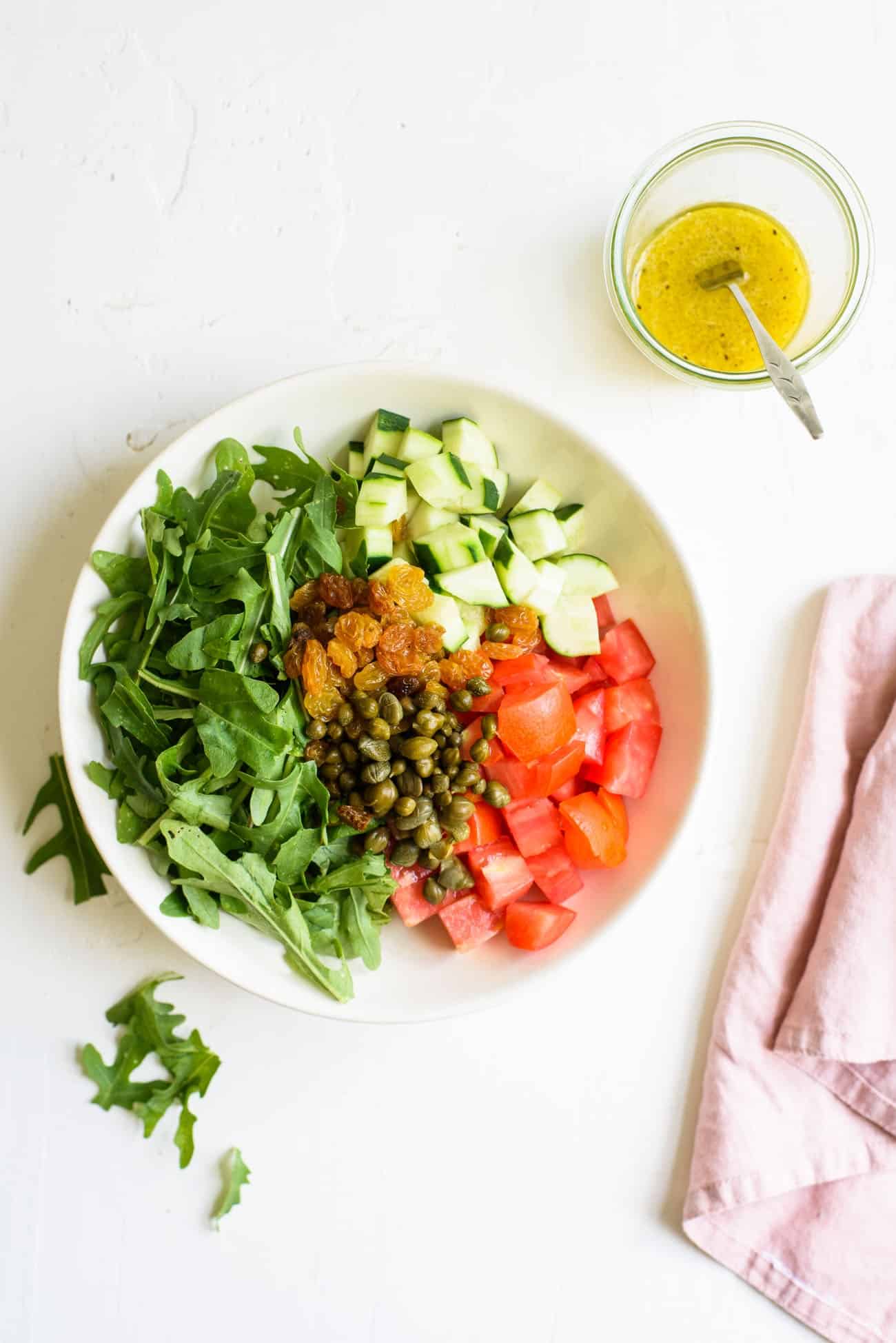Ingredients to make a summer farro salad in a white bowl: arugula, cucumbers, tomatoes, capers, and raisins