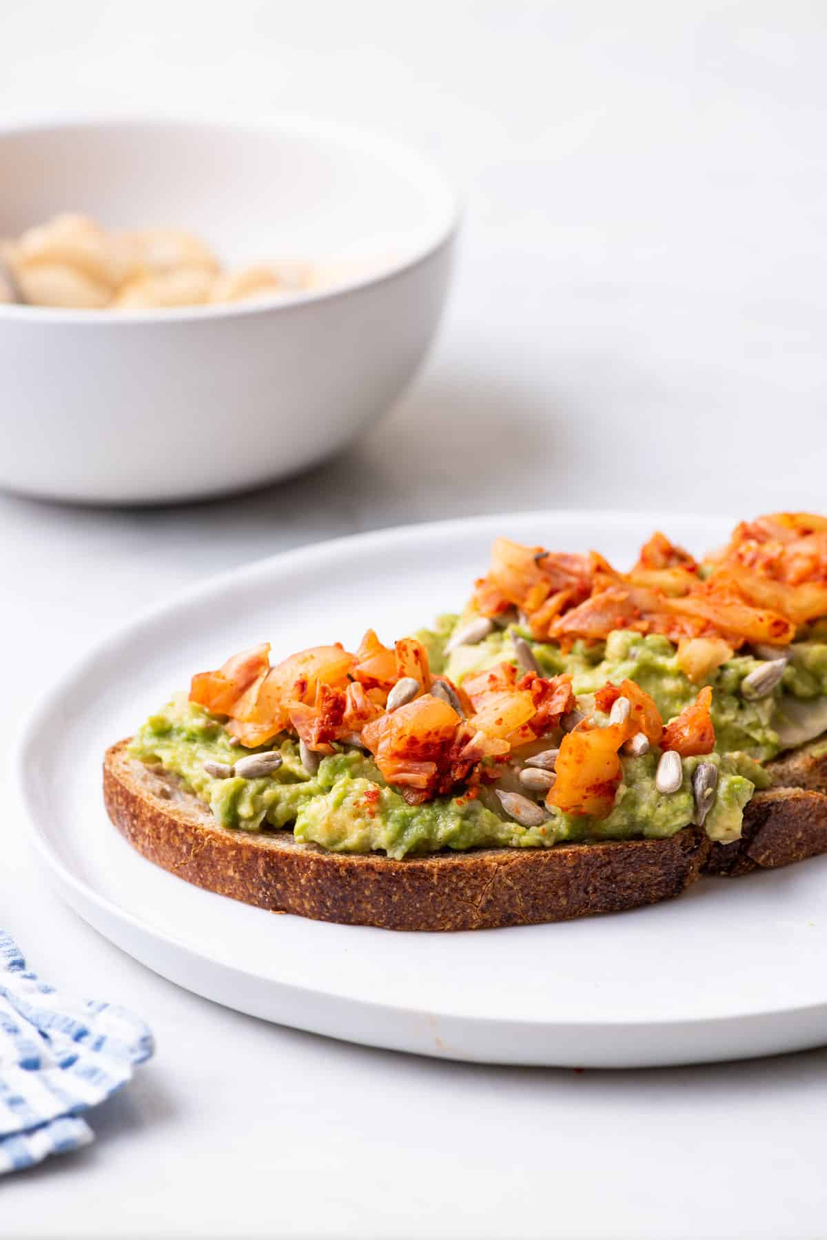 Avocado bean toast topped with sunflower seeds and kimchi, on a white.