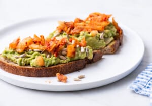 Avocado bean toast topped with sunflower seeds and kimchi on a white plate.