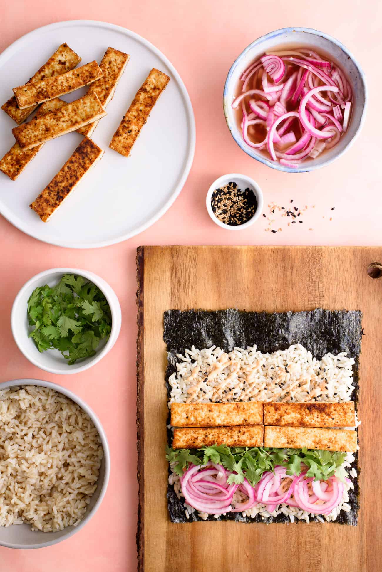 Assembly of sushi burrito recipe with nori on wooden cutting board surrounded by fillings in bowls