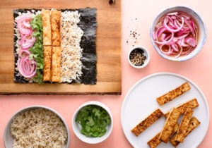 Assembly of sushi burrito recipe with nori on wooden cutting board surrounded by fillings in bowls