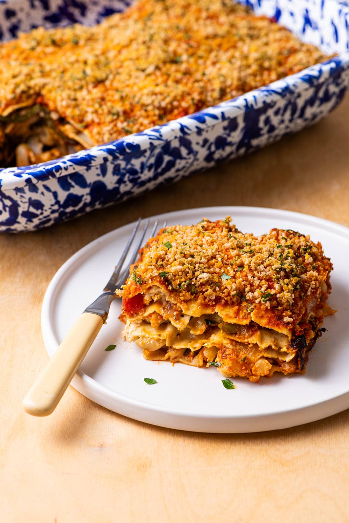 A vegan vegetable lasagna in a blue speckled casserole dish on a wooden table with white wine and a stack of plates.