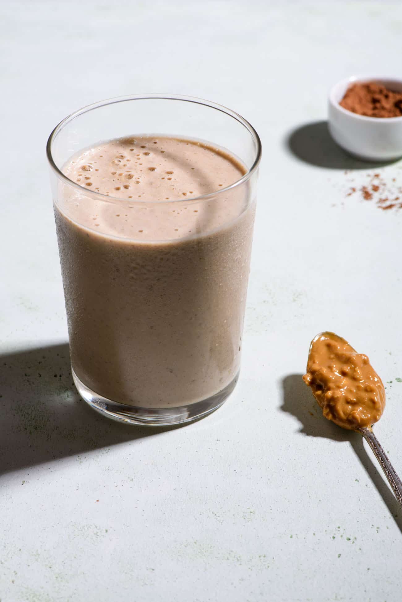 Healthy peanut butter banana smoothie in a glass next to spoon with peanut butter and cocoa powder