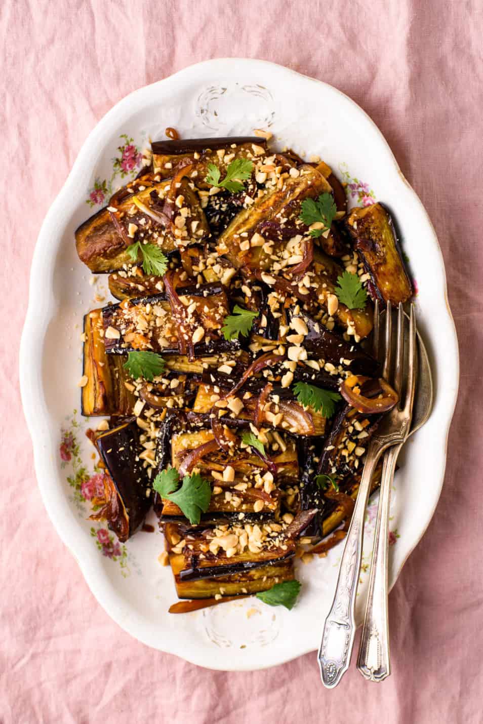 Soy-glazed pan-fried eggplant sprinkled with crushed peanuts and cilantro on a vintage oval platter on a pink tablecloth
