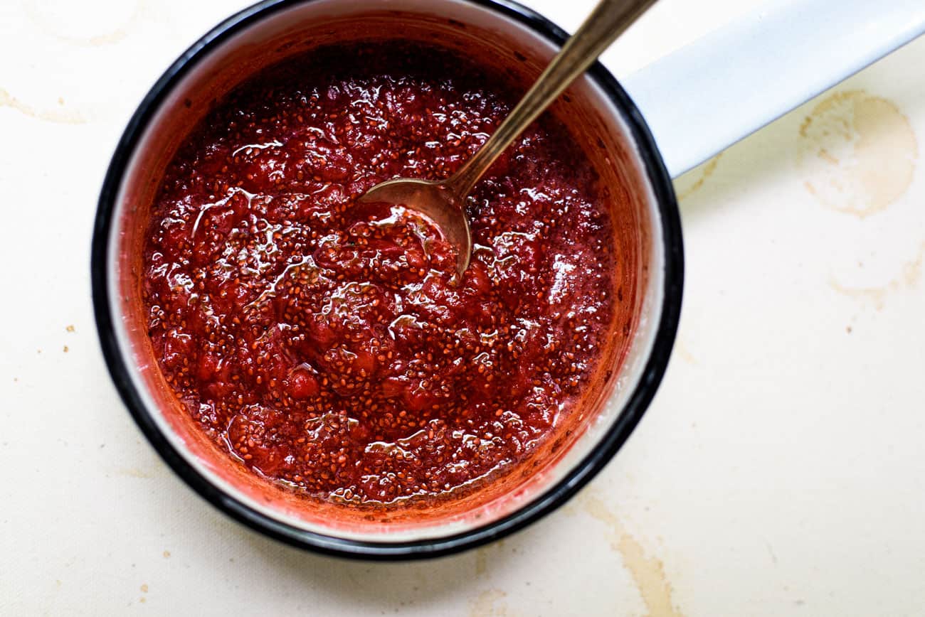 Strawberry chia jam in a small white pot with a metal spoon on a beige tablecloth