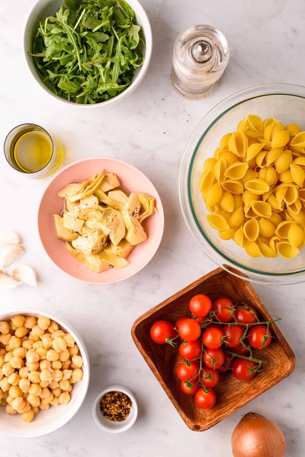 Ingredients gathered to make a no-mayo summer pasta salad: olive oil, arugula, pasta, marinated artichokes, cherry tomatoes, and chickpeas