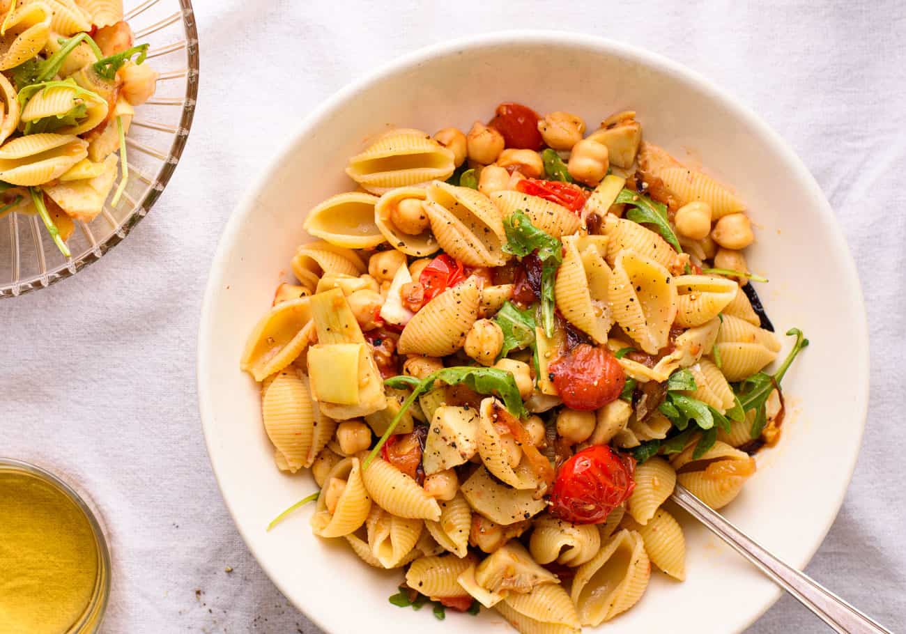 Summer pasta salad with tomatoes, artichokes, chickpeas, and arugula in a white bowl on a white tablecloth