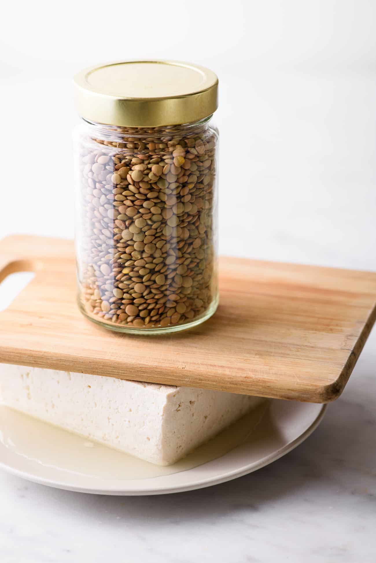 Tofu on a white plate being weighed down by a small wooden board and a jar of lentils - how to press tofu without a tofu press