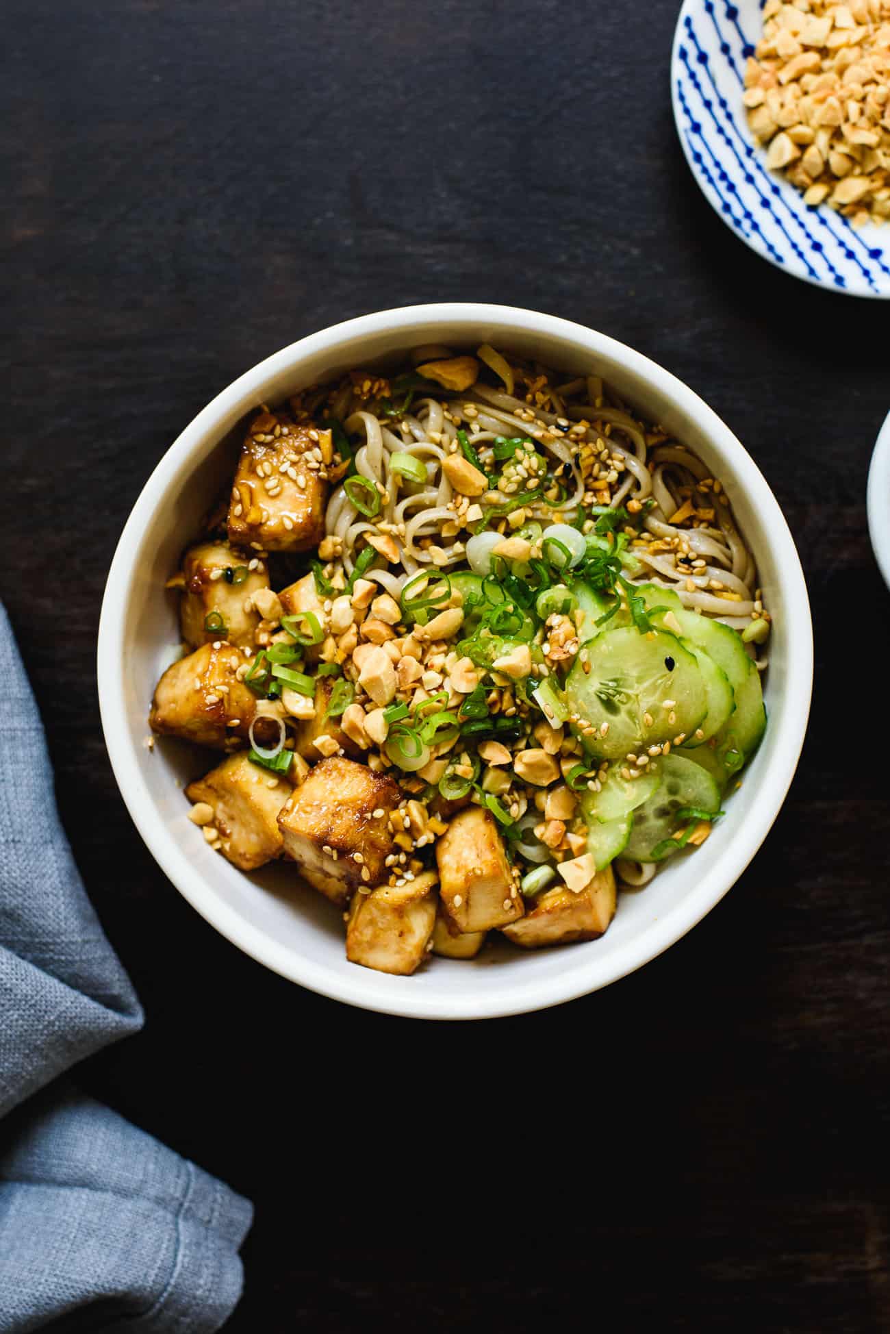 Soba noodle bowl with cucumbers, scallions, peanuts, and baked tofu on a dark wooden table