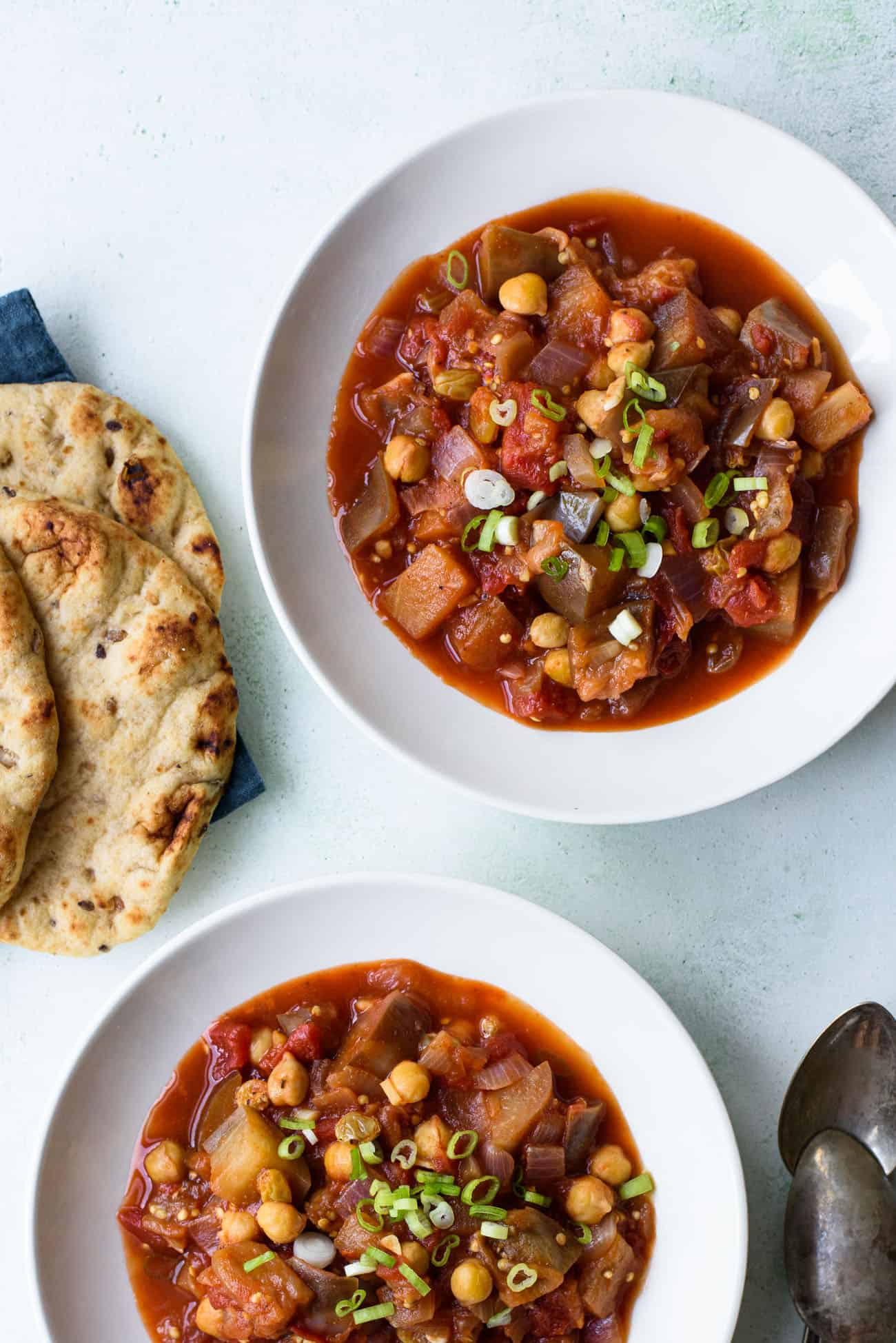 Two white bowls of eggplant chickpea stew next to a pile of flatbreads