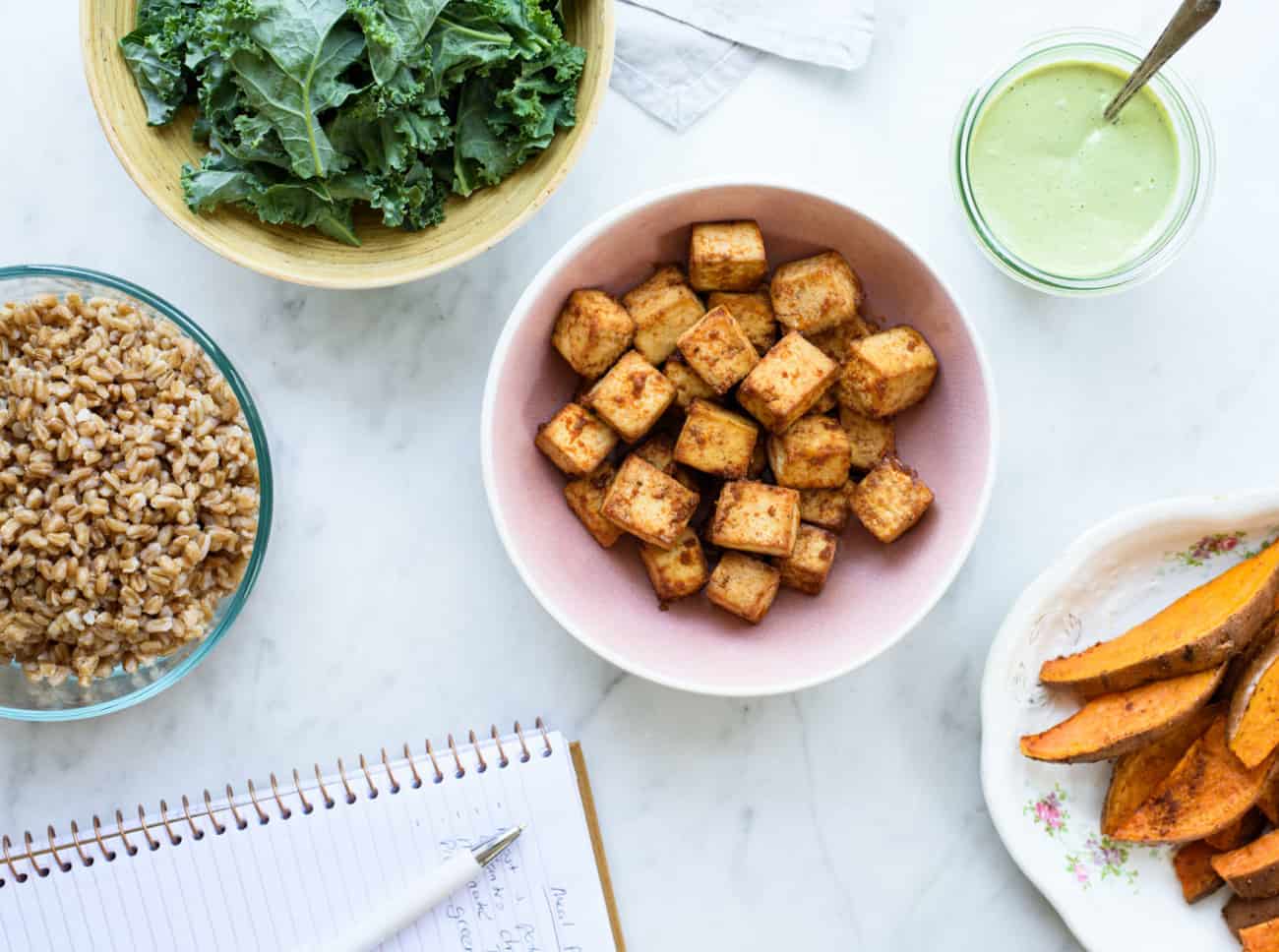 Completed plant-based meal prep: roasted sweet potatoes, baked tofu, creamy cilantro dressing, and farro, laid out on a marble table next to a notebook