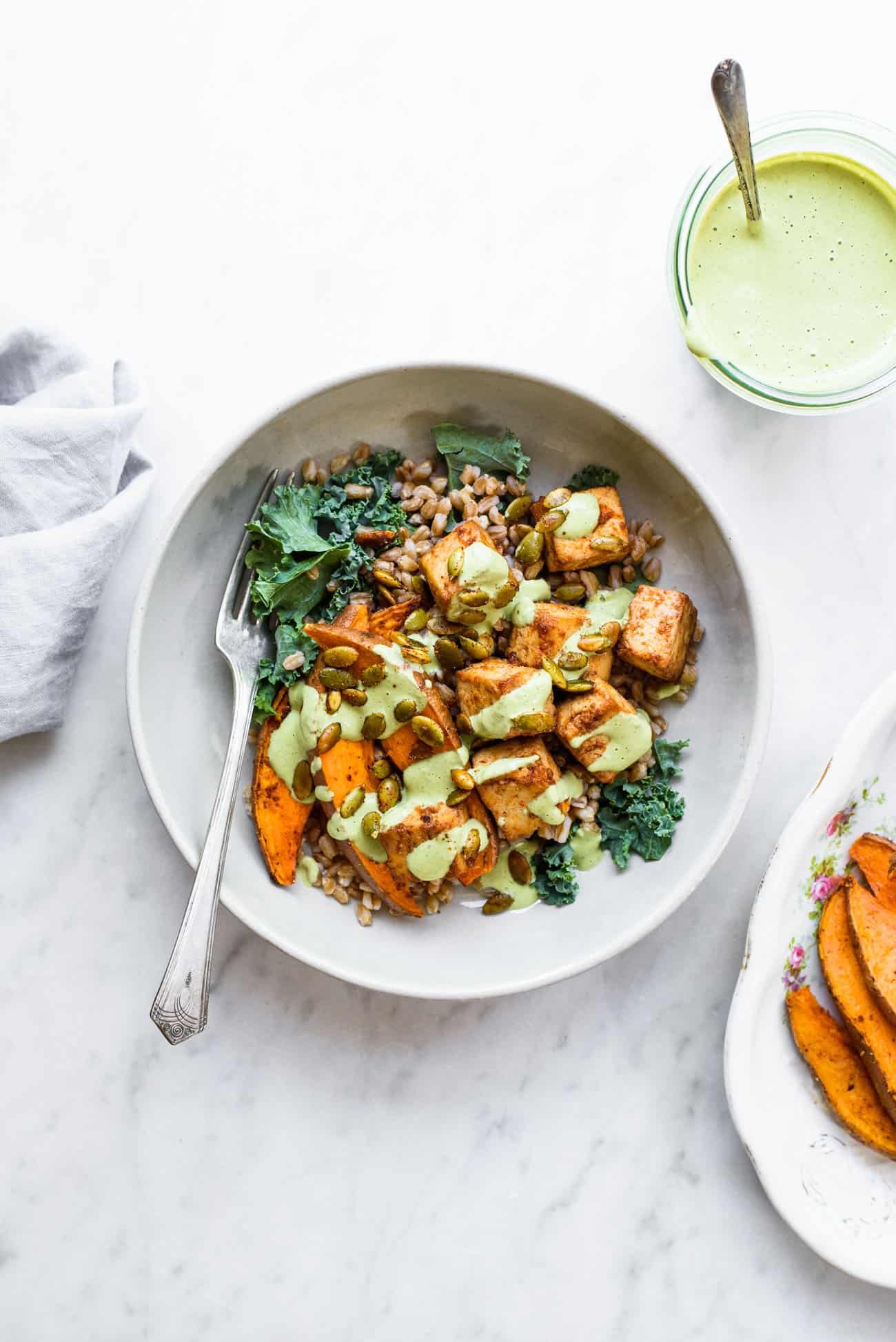 A bowl of farro, tofu, sweet potatoes, and kale with cilantro tahini dressing on a marble table