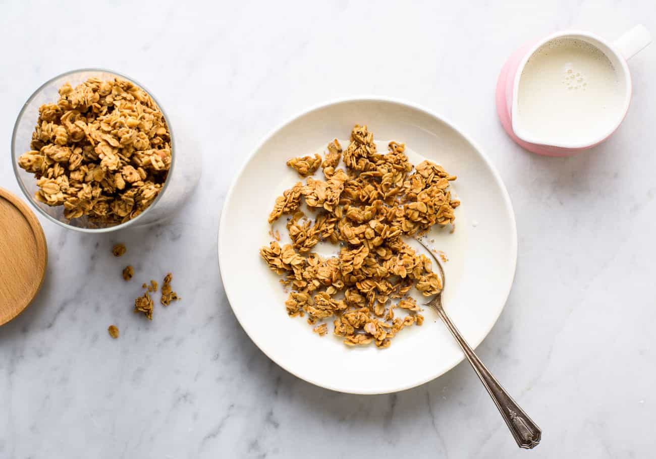 Peanut butter granola in a white bowl on a marble table next to a pink milk pourer