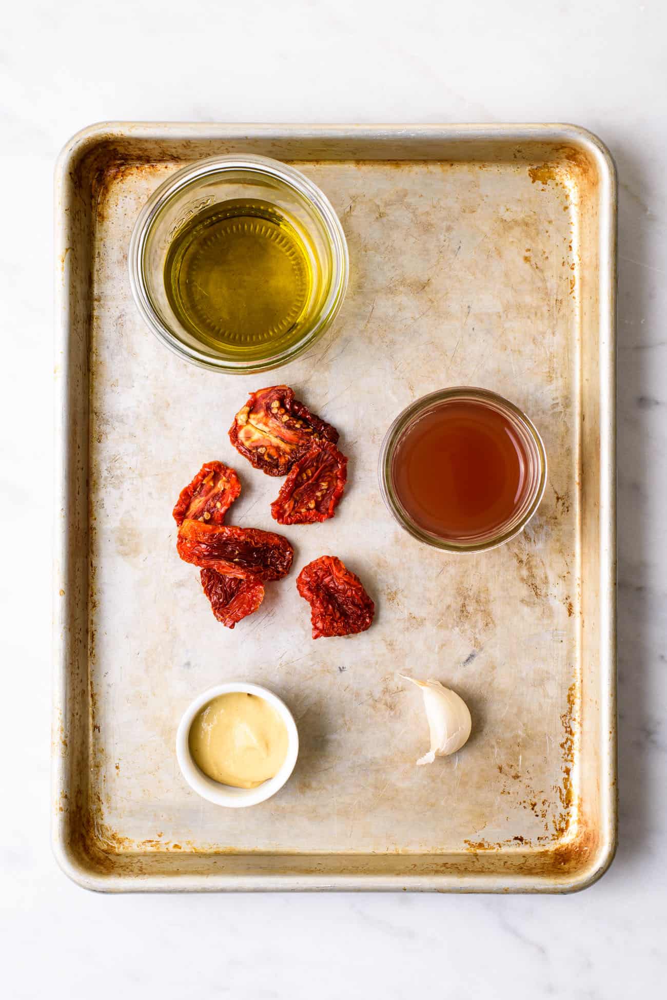 Ingredients to make sun-dried tomato vinaigrette gathered on a baking sheet: olive oil, sun-dried tomatoes, vinegar, mustard, and garlic