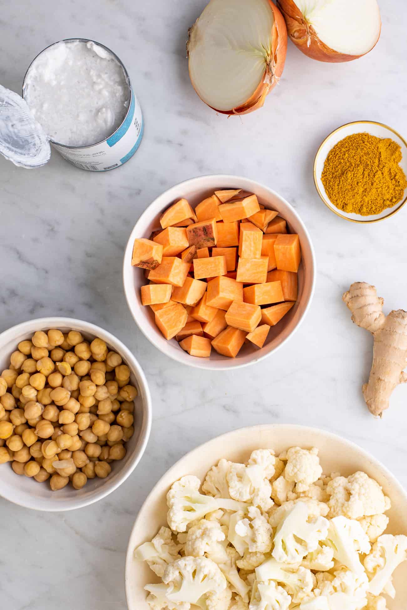 Ingredients to make cauliflower rice curry gathered on a marble table: onion, coconut milk, sweet potatoes, curry powder, chickpeas, cauliflower