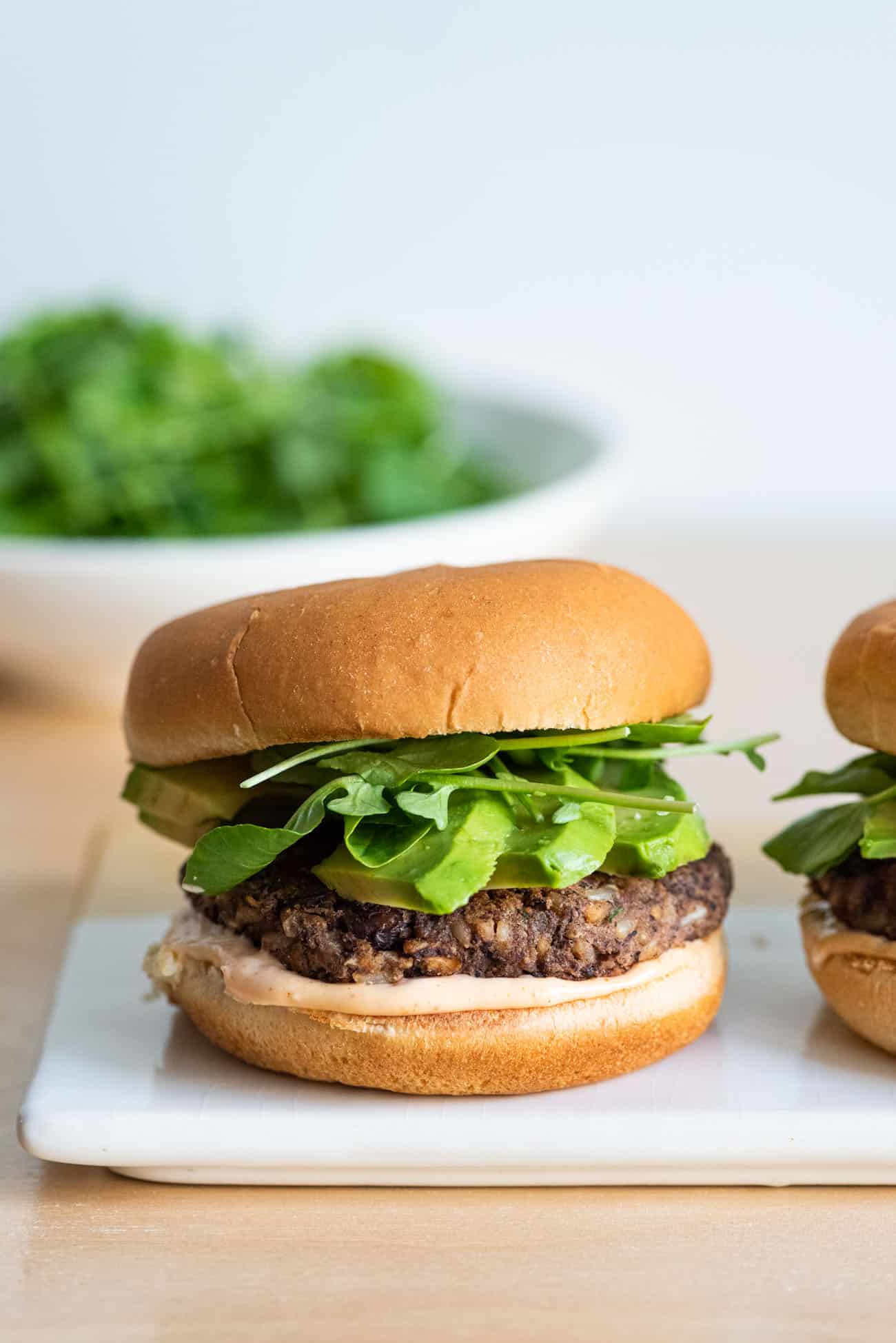 A vegan black bean burger with avocado, watercress, and chipotle mayo on a ceramic tray with a bowl of greens in the background