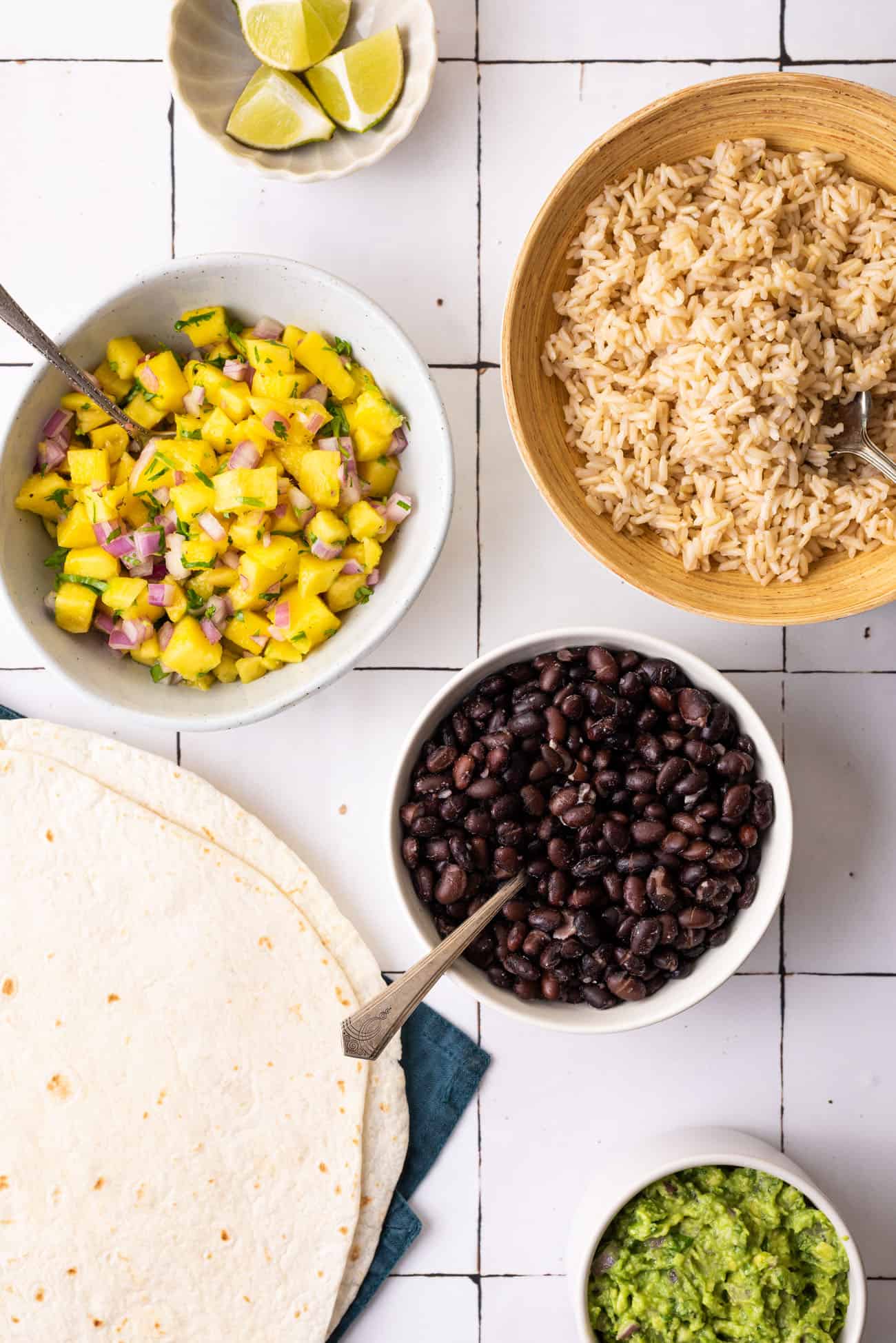 Ingredients to make vegan black bean burritos gathered on a white-tiled table: brown rice, black beans, mango salsa, guacamole