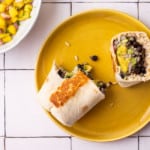 Vegan black bean burrito with mango salsa, cut in half, on a yellow plate on a white-tiled table