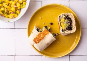 Vegan black bean burrito with mango salsa, cut in half, on a yellow plate on a white-tiled table