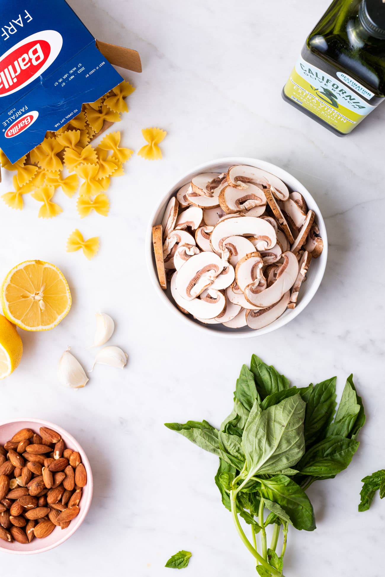 Ingredients gathered on a marble counter: Barilla farfalle, olive oil, mushrooms, garlic, almonds, and basil.