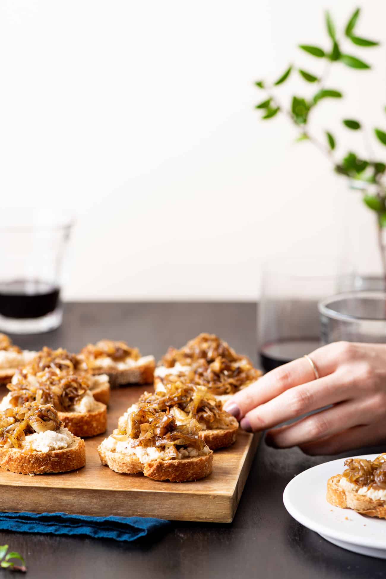 Woman's hand reaching for caramelized onion crostini with vegan cashew cheese.