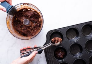 Woman's hands scooping brownie dough into a muffin tin from a food processor bowl
