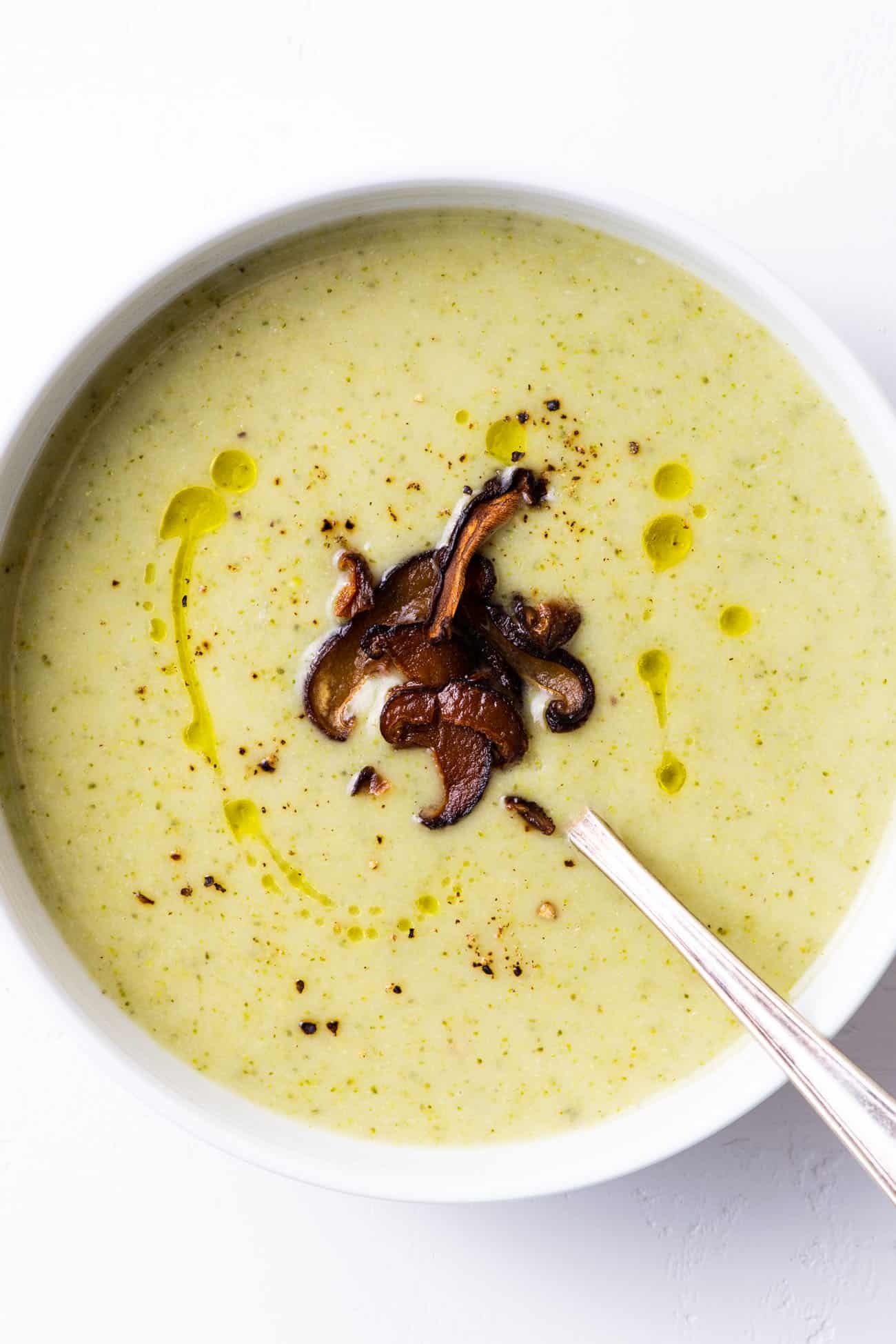 Dairy-free broccoli potato soup garnished with shiitake 'bacon', olive oil, and black pepper, in a white bowl.