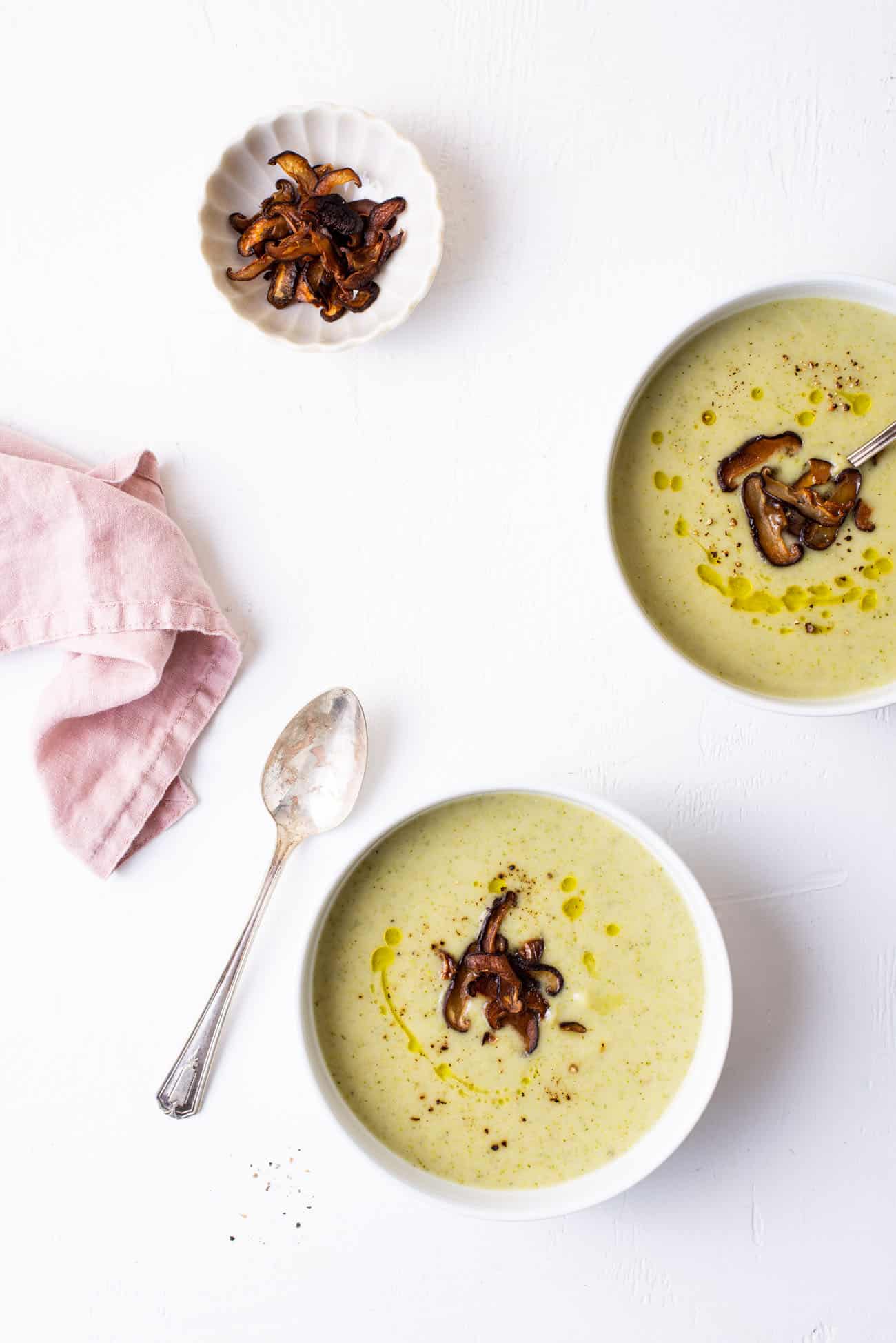 2 bowls of creamy broccoli potato soup, garnished with mushroom bacon, on a white table.