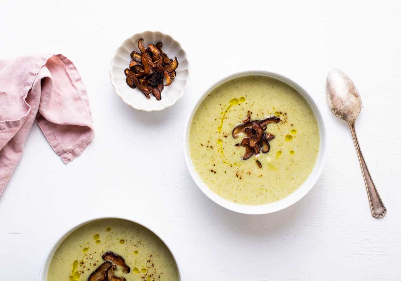 2 bowls of creamy broccoli-potato soup, garnished with mushroom bacon, on a white table