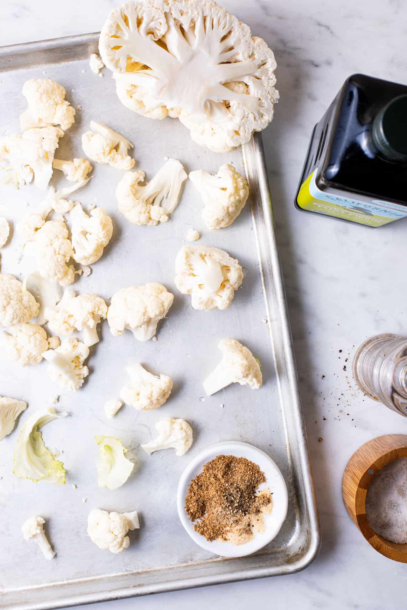 Raw cauliflower on a baking sheet next to olive oil and spices.