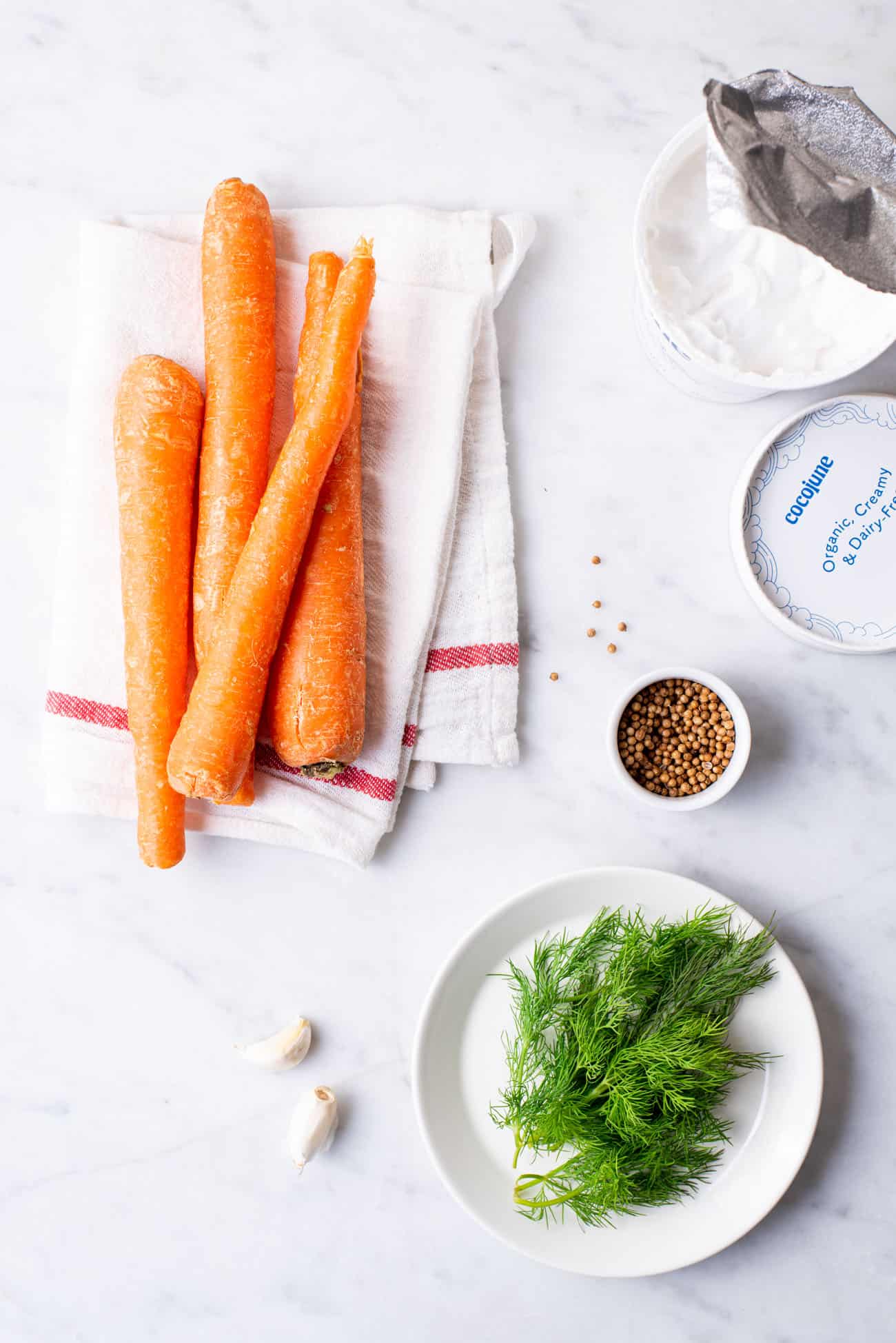 Carrots, vegan yogurt, coriander seeds, garlic cloves, and dill, gathered on a marble table