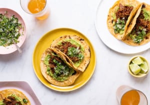 Vegan carnitas tacos on various plates, next to glasses of beer and cilantro-onion relish