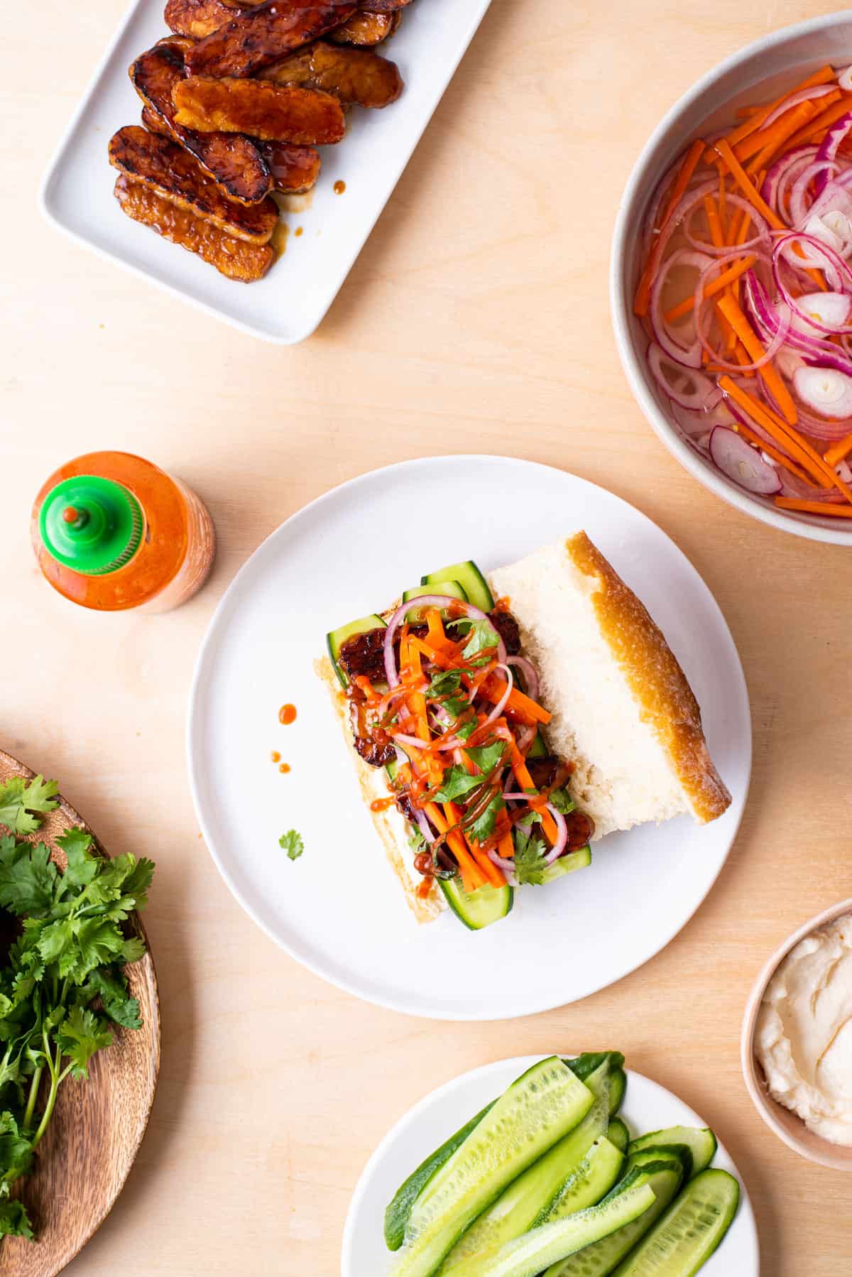 Assembling a tempeh banh mi sandwich: glazed tempeh, pickled vegetables, cucumbers, and cilantro, laid out on a wooden table.