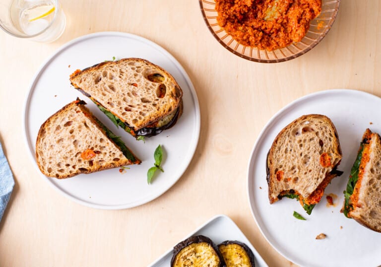 Roasted eggplant sandwiches with romesco sauce on white plates on a wooden table.