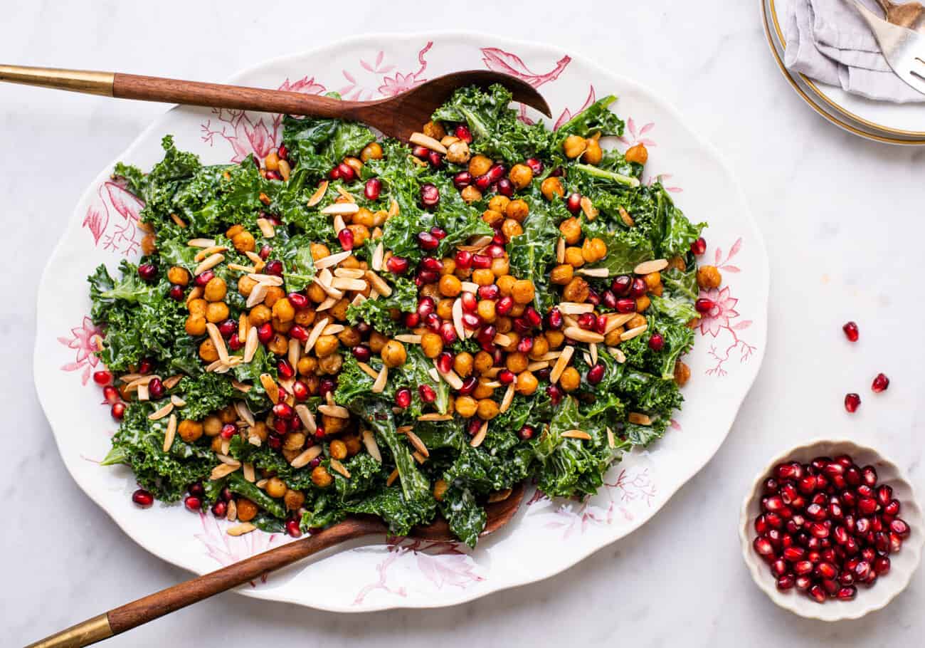 Massaged kale salad with tahini dressing and crispy chickpeas on an oval platter next to pomegranate seeds.