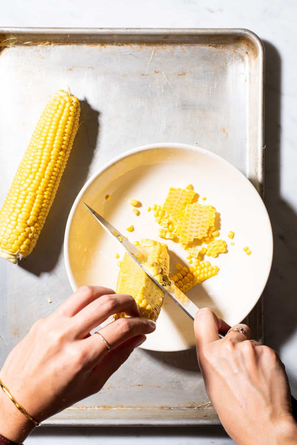 Woman's hands cutting corn kernels off the cob in a beige bowl.