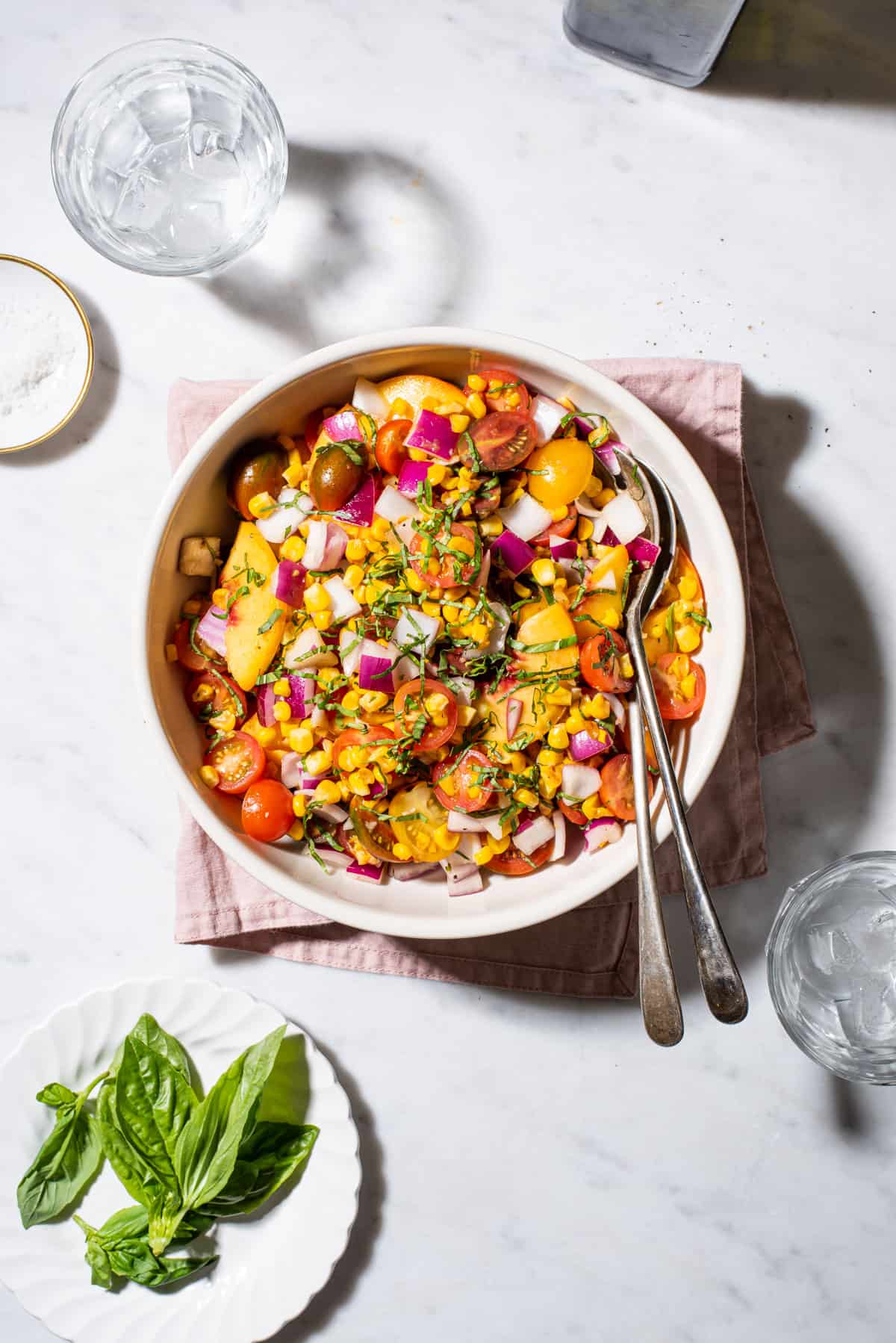 Bowl of tomato peach salad with corn next to a pile of basil on a marble table.