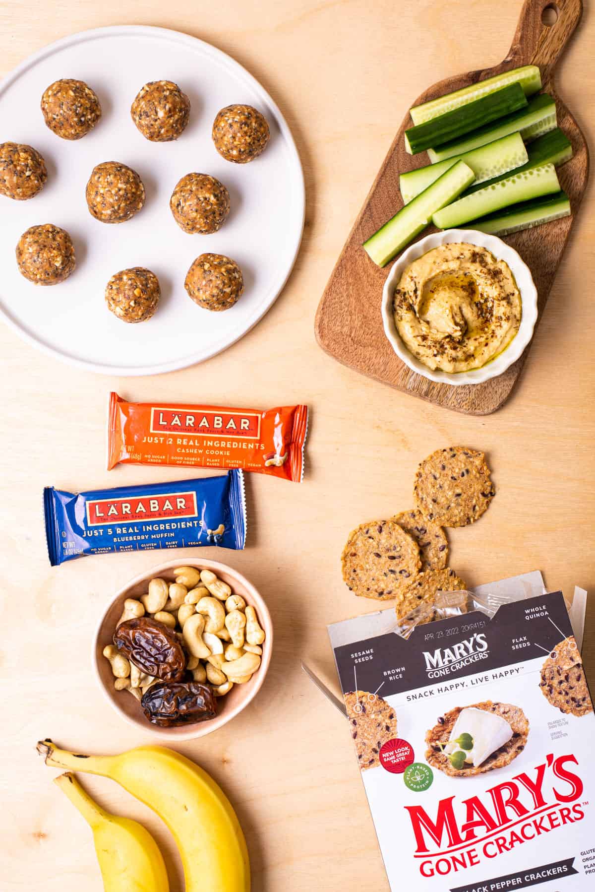 Plant-based healthy snacking ideas gathered on a wooden table: including energy balls, hummus, Larabars, and bananas.