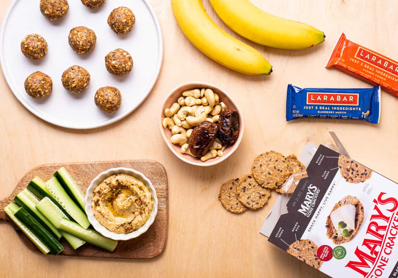 Healthy snacks on a wooden table, including energy balls, hummus, nuts, and bananas.