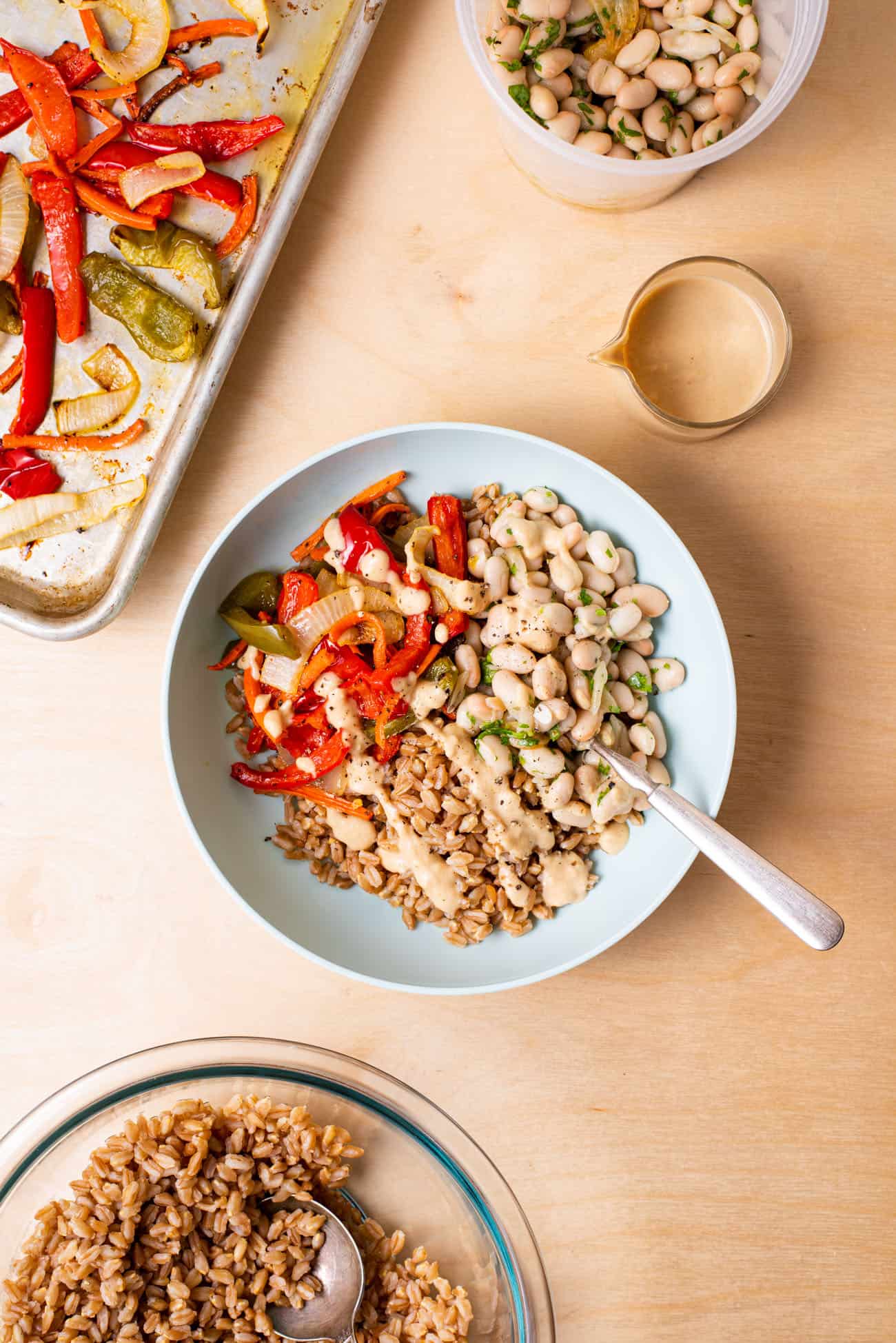 Farro bowl next to a tray of roasted vegetables, white bean salad, and tahini dressing.