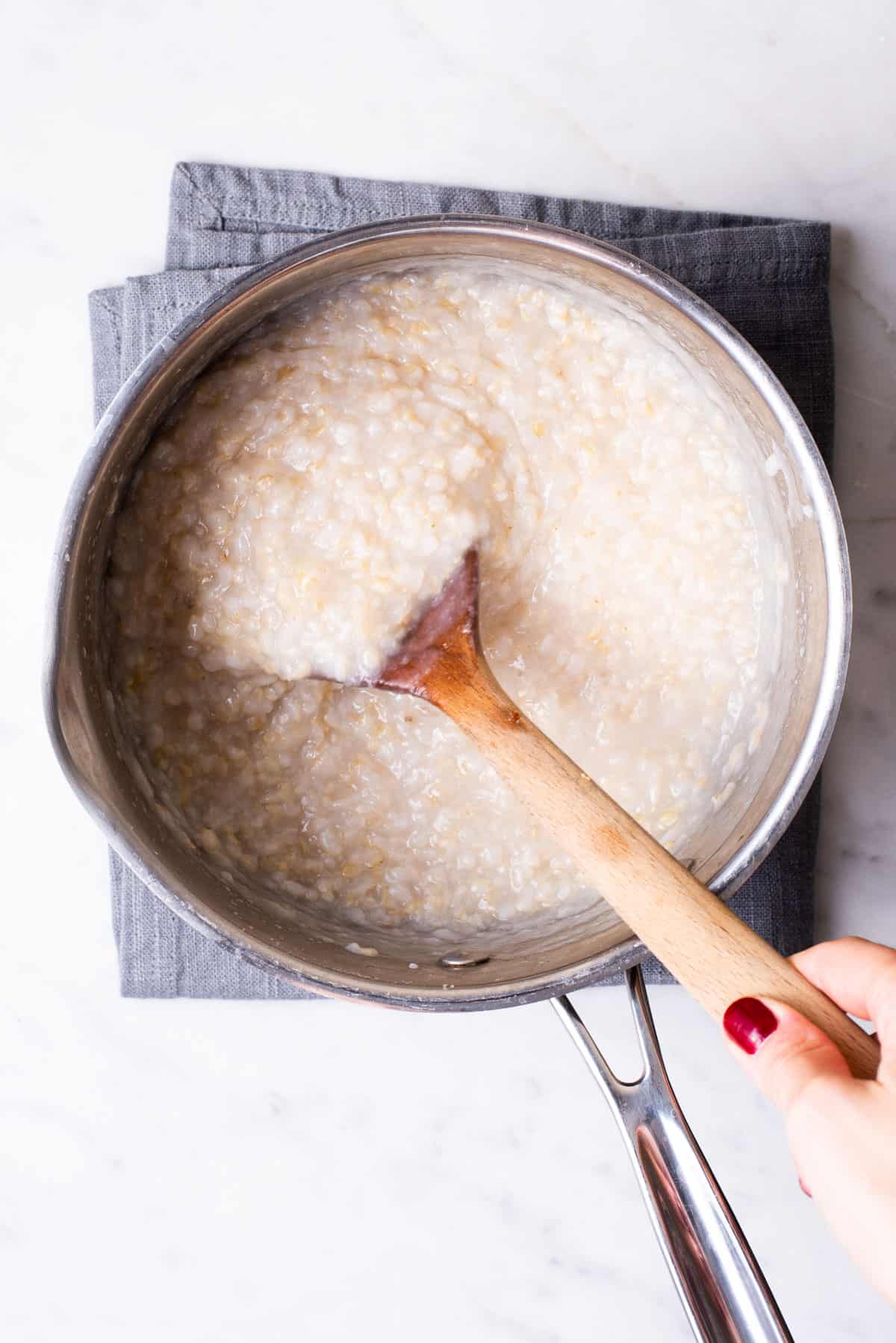 Woman's hand stirring brown rice congee in a pot with a wooden spoon.